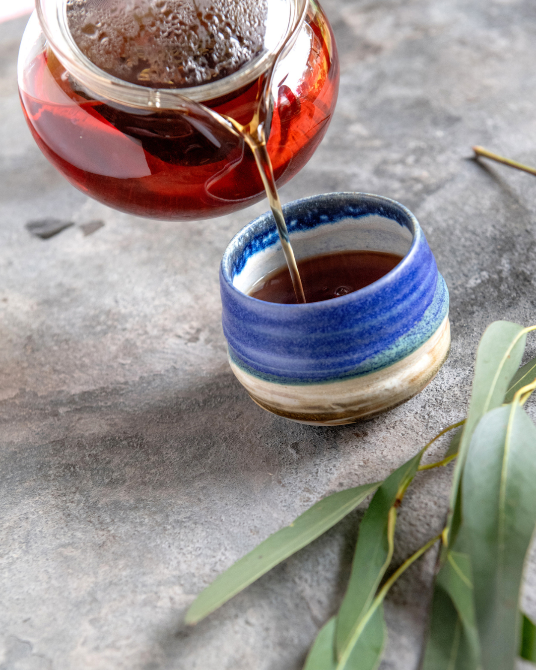 glass tea pot of tea being poured into a pottery mug