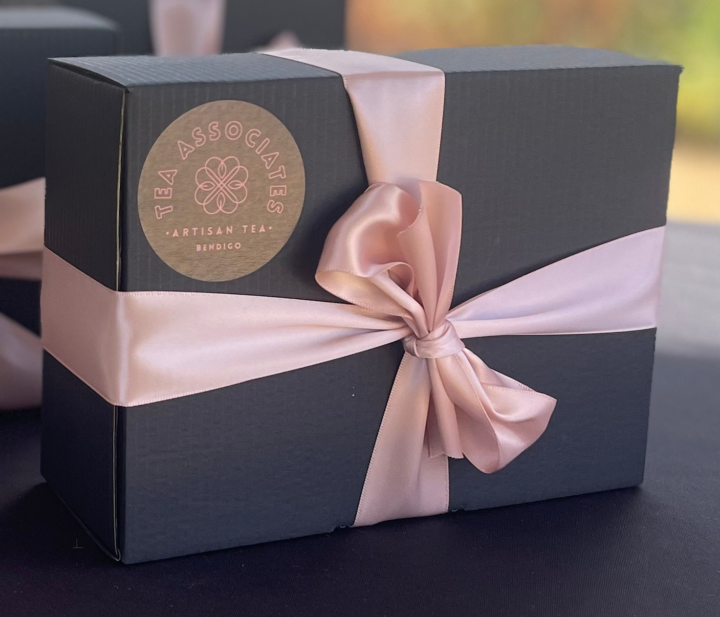A black gift box with a pink ribbon bow and a tag that reads 'A Year of Tea' by The Tea Associates.